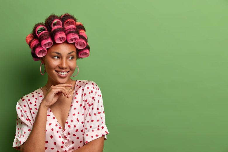 portrait-beautiful-smiling-woman-keeps-hand-chin-poses-with-hair-rollers-head-perfect-curls-dressed-nightwear-prepares-special-meeting-isolated-green-wall-empty-space_273609-39502